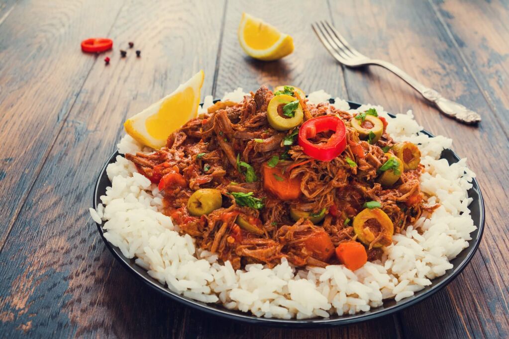 The national dish of Cuba, Ropa Vieja, tender shredded beef stewed with vegetables and spices on a black plate with a boiled rice on the wooden table.
