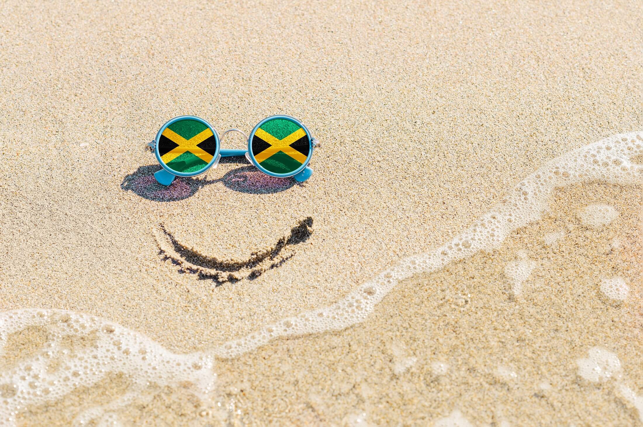 A painted smile on the sand and sunglasses with the flag of Jamaica. The concept of a positive and successful holiday in the resort of Jamaica.