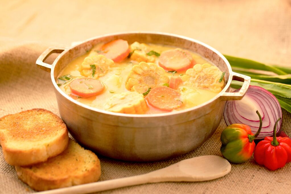 big pot of caribbean corn soup with corn, carrots, bread, pepper, onion, wooden pot spoon, shadow beni, chive, seasoning