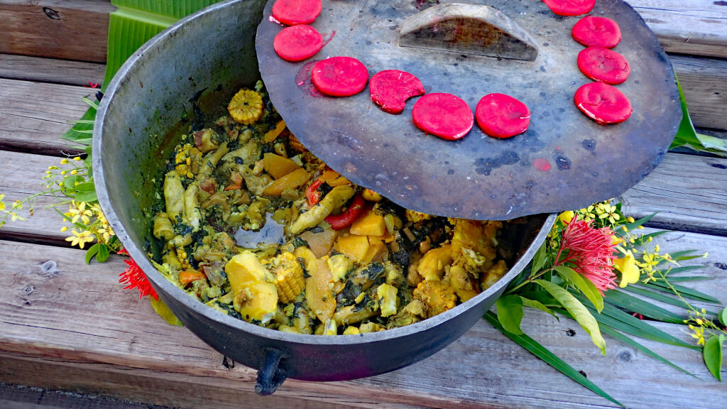Large, rustic iron pot full of bright yellow Caribbean curry stew of corn on the cob, saltfish, dumplings and spices, adorned by colorful, tropical flowers and palms from a Grenada oil down festival.
